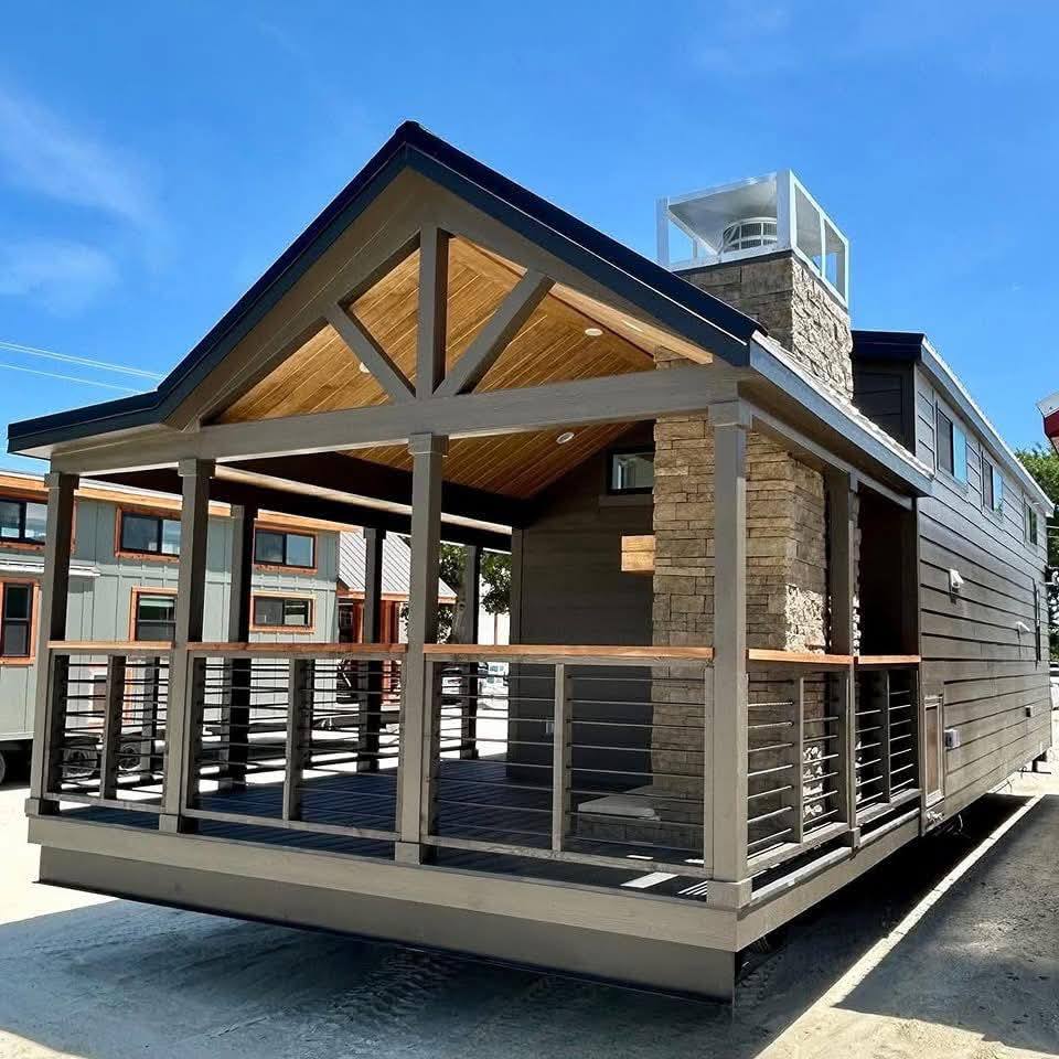 A modern tiny house with a wooden porch and stone chimney, featuring gray siding and metal railings, stands under a clear blue sky in a sunny setting.