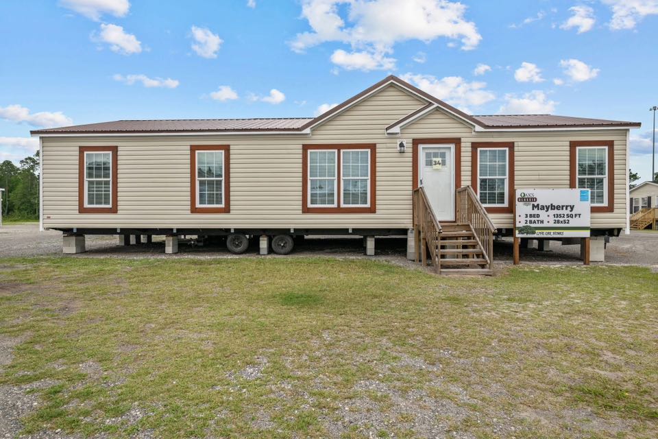 Tan mobile home with brown trim on a grassy lot. A wooden staircase leads to the entrance; a sign reads "Mayberry, 3 Bed, 2 Bath, 1920 SQ FT." Clear sky above.