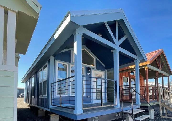 Blue tiny home with modern design features a gable roof, large windows, and a small porch with railings. It exudes a cozy and inviting feel under a clear sky.