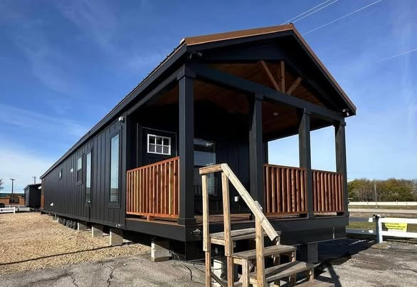 Black tiny home on stilts with a small wooden porch, surrounded by a gravel lot under a clear blue sky. Wooden steps lead up to the entrance.