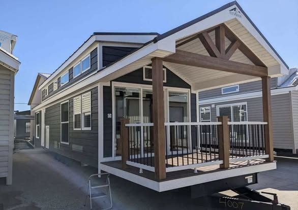 Modern tiny house with dark siding and a peaked roof, featuring a wooden porch with white railings. Other tiny homes are visible in the bright background.