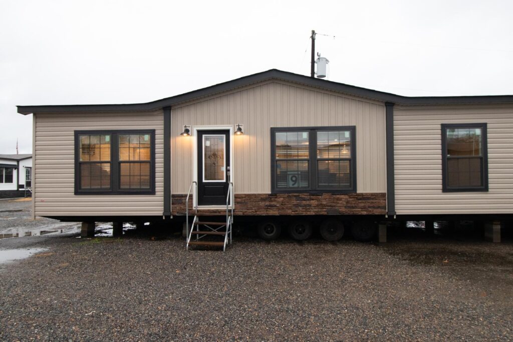 A beige mobile home with large windows and a central door on a gravel lot. Lit porch lights add warmth on a cloudy day, creating a cozy atmosphere.