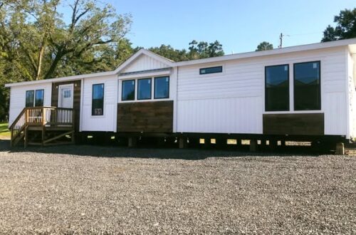 A modern, white mobile home with brown accents sits on a gravel lot. It features large windows, a small porch, and trees in the background under a clear sky.