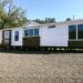A modern, white mobile home with brown accents sits on a gravel lot. It features large windows, a small porch, and trees in the background under a clear sky.