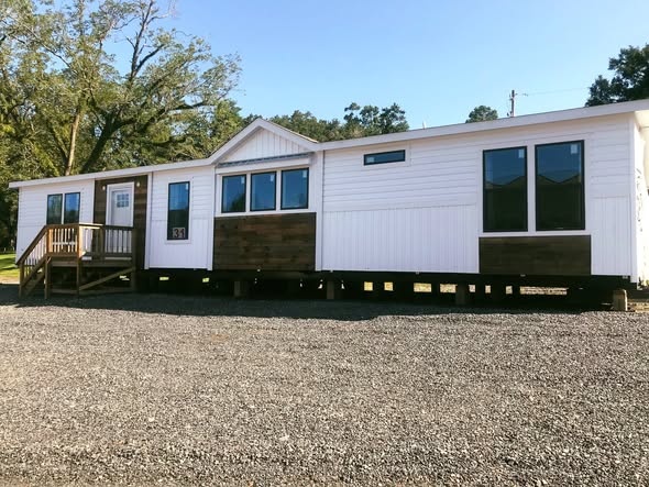 A modern, white mobile home with brown accents sits on a gravel lot. It features large windows, a small porch, and trees in the background under a clear sky.