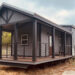A modern tiny house on wheels with a dark wooden porch, railing, and gabled roof. It stands in a grassy area under a cloudy sky, conveying a rustic charm.