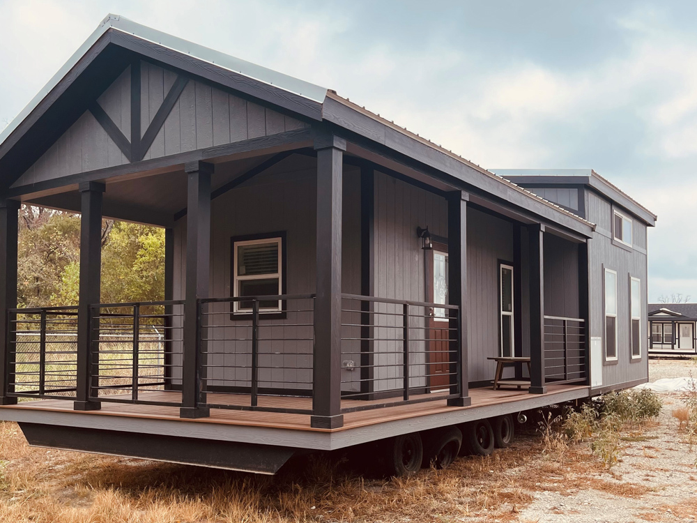 A modern tiny house on wheels with a dark wooden porch, railing, and gabled roof. It stands in a grassy area under a cloudy sky, conveying a rustic charm.