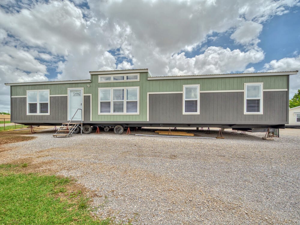 A modern, gray and green manufactured home on a gravel lot beneath a partly cloudy sky. The home has large windows and a small metal staircase.