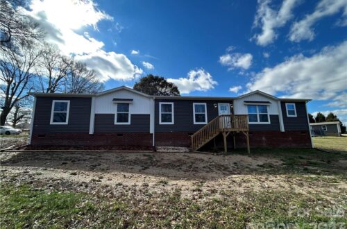 Single-story house with dark siding and white trim, featuring a small wooden porch. The sky is blue with scattered clouds, creating a calm rural setting.