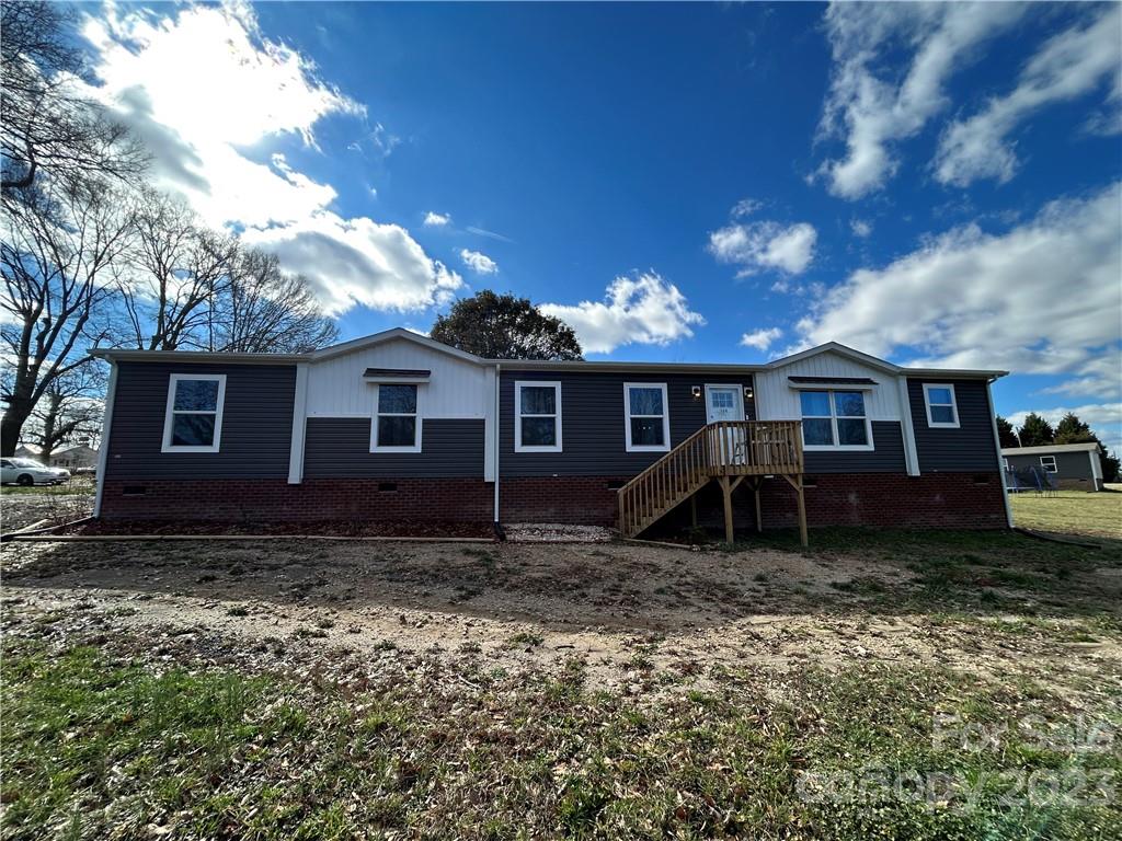 Single-story house with dark siding and white trim, featuring a small wooden porch. The sky is blue with scattered clouds, creating a calm rural setting.