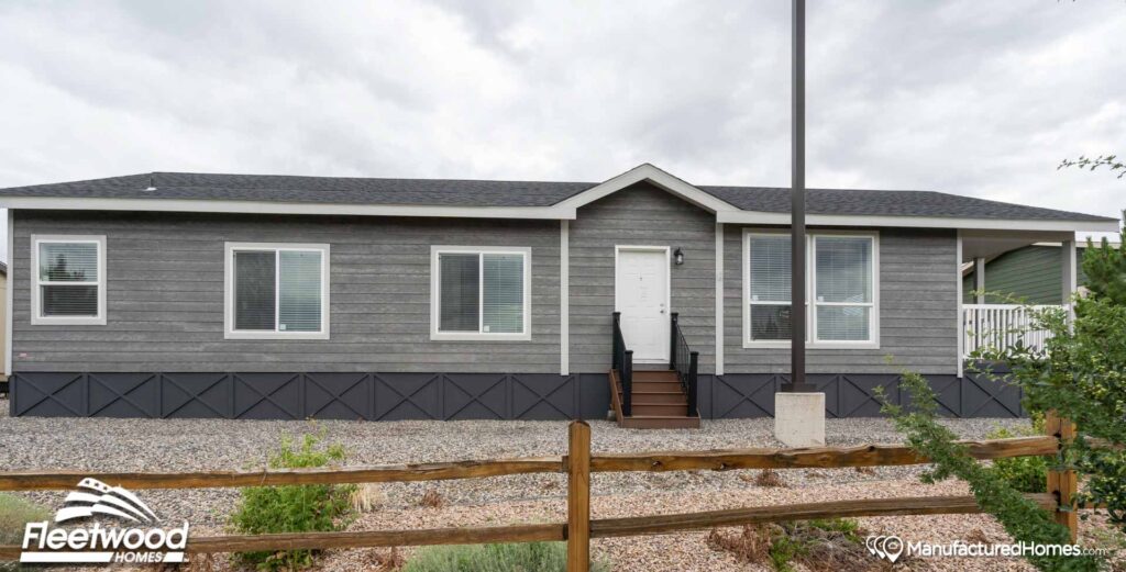 Single-story manufactured home with gray siding, white trim, and a central entrance. Surrounded by gravel landscaping and a wooden fence, under a cloudy sky.
