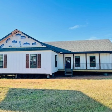 A white ranch-style house with a front porch, wooden shutters, and incomplete siding stands in a sunny yard, conveying a sense of potential and craftsmanship.