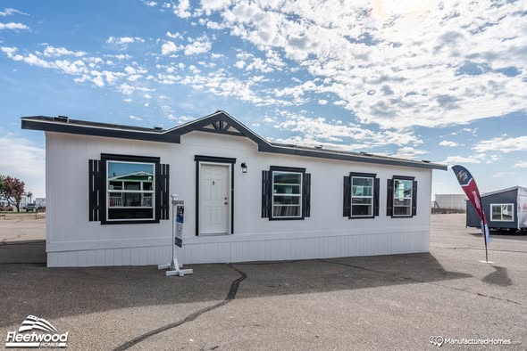 A white manufactured home with black shutters is set against a partly cloudy sky. A small flag and sign are in front, suggesting it's a display model.