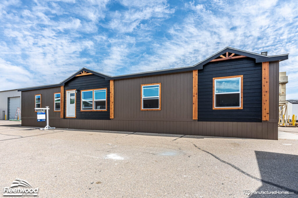 A modern manufactured home with dark brown and black panel siding sits on a lot under a blue sky with scattered clouds. The structure has multiple windows and a simple, clean design.