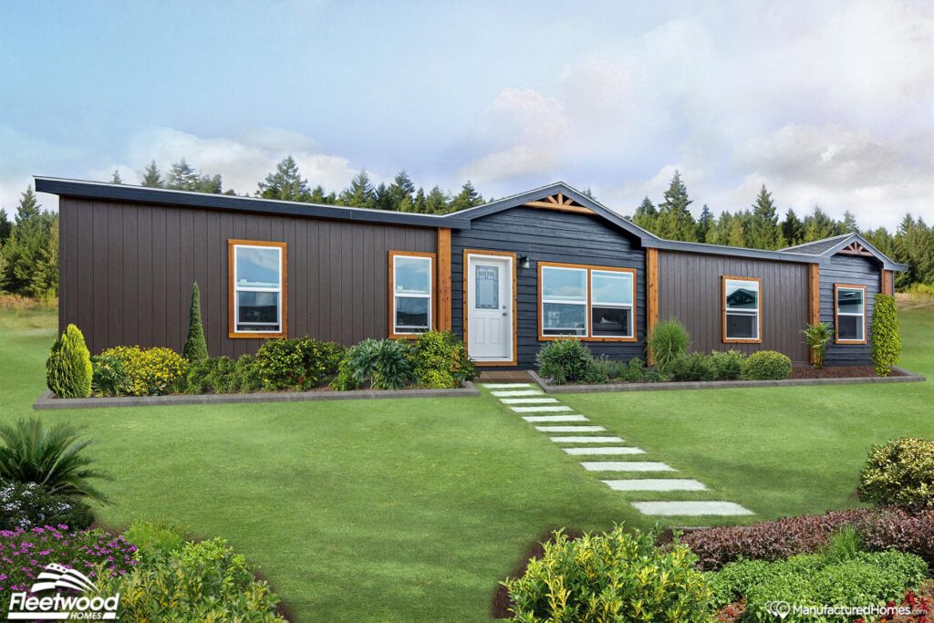A modern manufactured home with dark wood siding, white-framed windows, and a central white door. It's surrounded by manicured greenery and a stone path, set against a backdrop of lush trees and a partly cloudy sky.