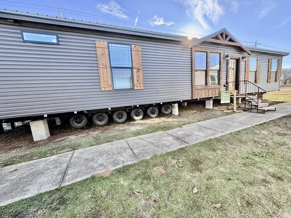 A modern gray mobile home with wooden shutters and a gabled front porch is elevated on concrete blocks. It’s set in a grassy area under a bright blue sky.