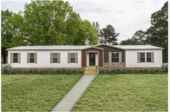 A white manufactured home with brown shutters sits amidst a lush green lawn. A concrete path leads to the front door. Trees are in the background.