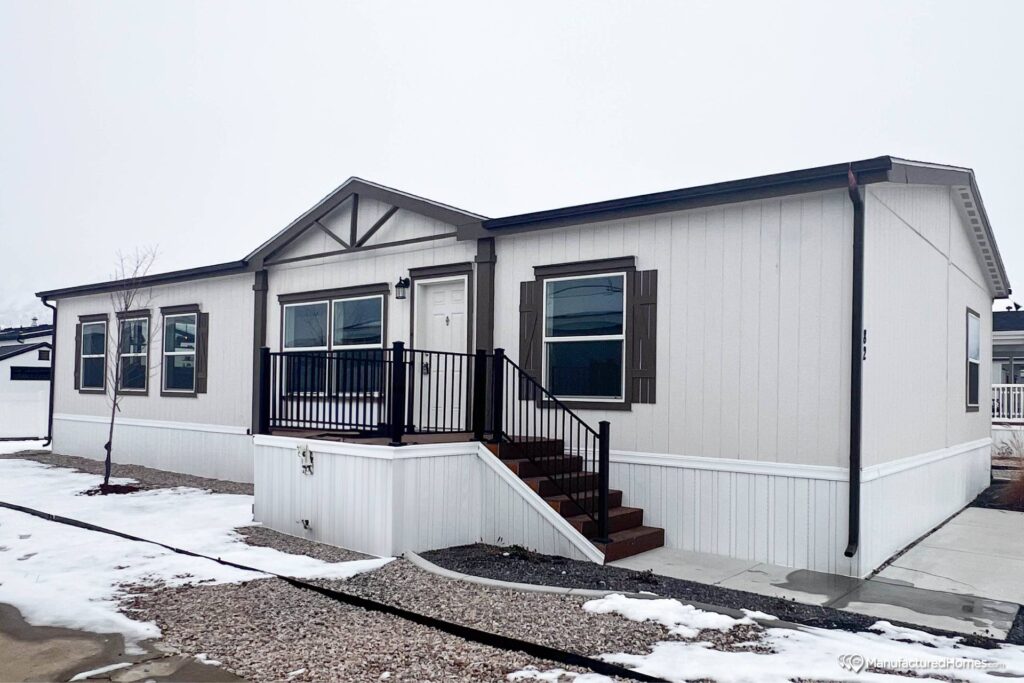 A single-story white manufactured home with dark trim, steps leading to a small porch, and three front windows. Snow and gravel are visible around the home.