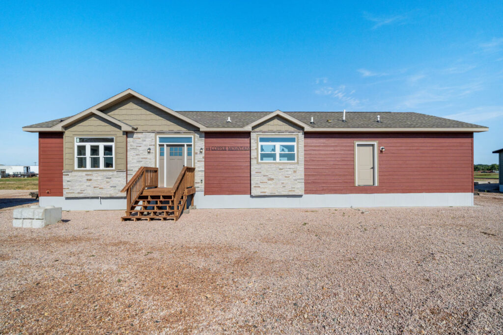 Single-story manufactured home with a red and beige exterior under a clear blue sky. Cement steps lead to the entrance, set on a gravel lot.
