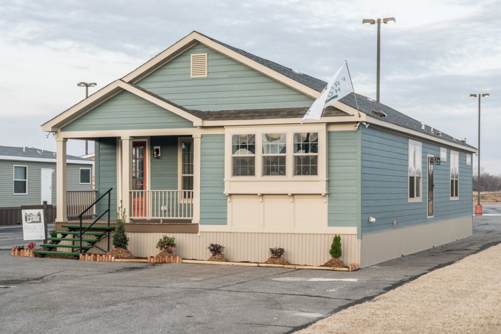 A cozy, blue-green modular home with a front porch and flag, set in an open lot. The home's welcoming facade and neat landscaping create a homely feel.