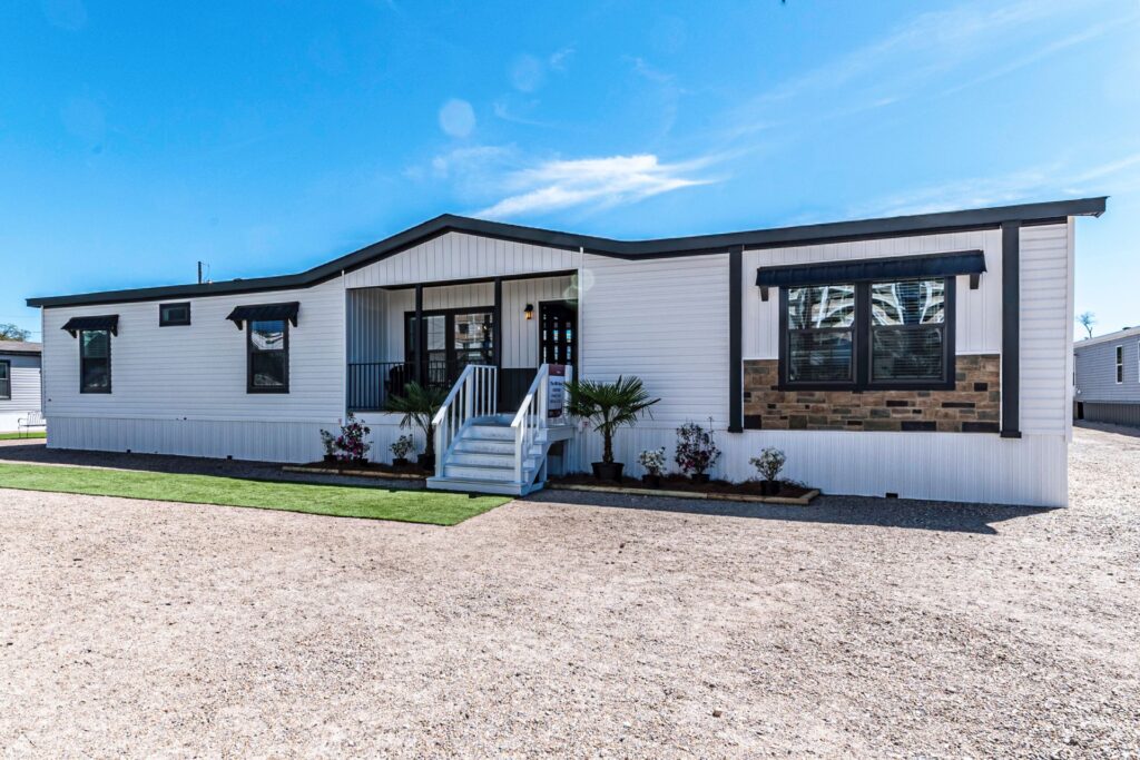A modern manufactured home with white siding, stone accents, and black trim under a clear blue sky. Features a small porch, stairs, and minimal landscaping.