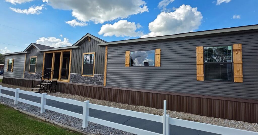 A modern manufactured home with dark gray siding and stone accents, wooden shutters on large windows, under a bright blue sky with clouds, alongside a white fence.