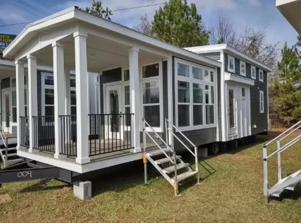 Elegant tiny house with large windows and a covered porch on a sunny day, set on a grassy area with a backdrop of trees, conveying coziness.