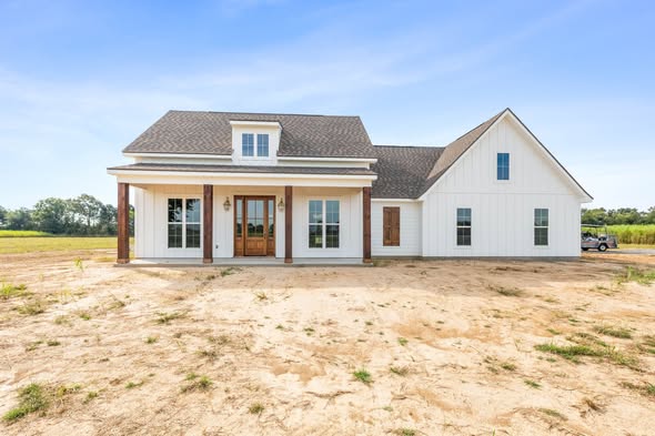 A newly built farmhouse-style home with white siding, a covered porch, and a gabled roof sits on a dirt lot under a clear blue sky, evoking tranquility.