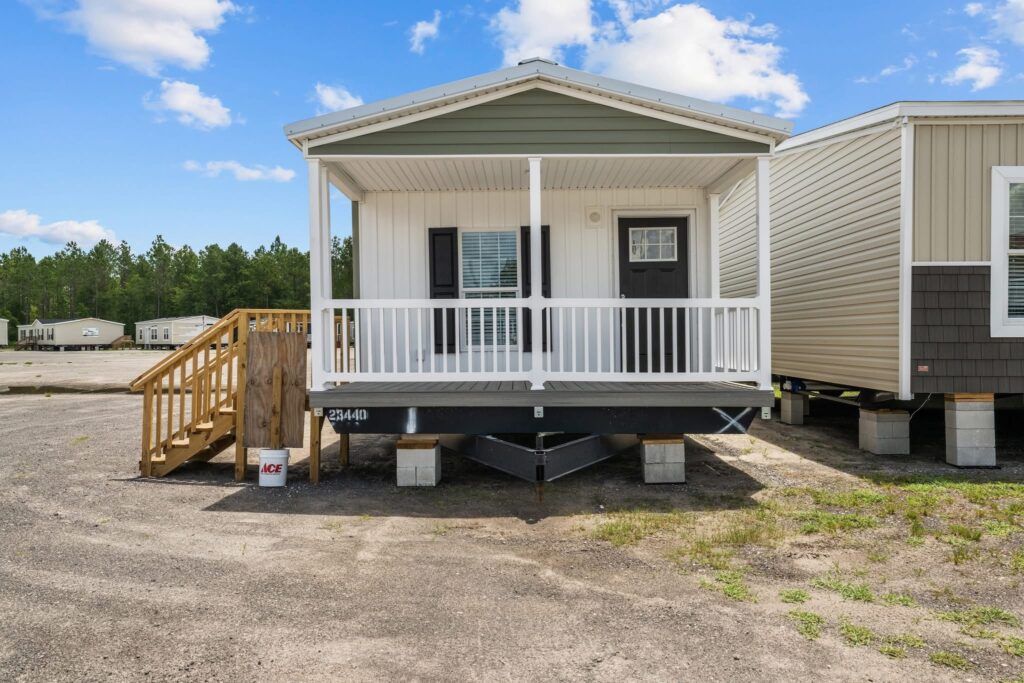 Small portable home on a gravel lot under a blue sky with clouds. It has a front porch, wooden steps, and is surrounded by other similar homes.