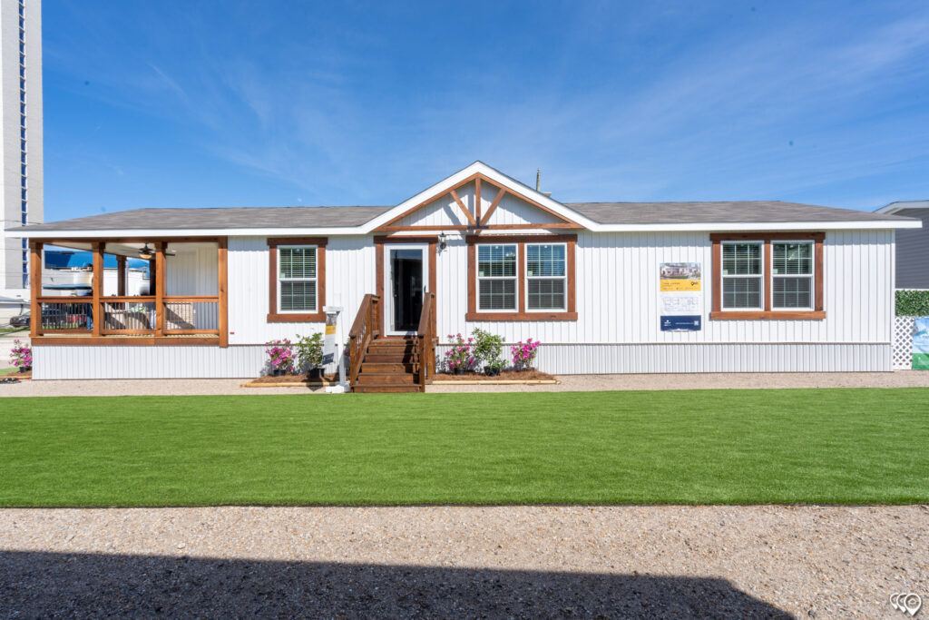 A white modular home with brown trim and front steps sits under a blue sky. It features several large windows and a porch to the left, surrounded by green grass and pink flowers, creating a welcoming atmosphere.