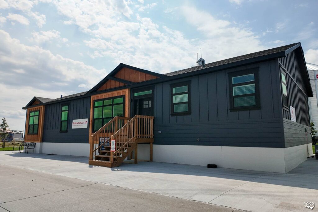 A modern, dark gray manufactured home with green-trimmed windows and a wooden staircase under a partly cloudy sky, conveying a welcoming tone.