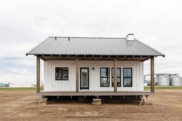 A small, white, wooden house with a gray metal roof sits on an open dirt lot. It features a porch with wooden posts. Silos are visible in the background.