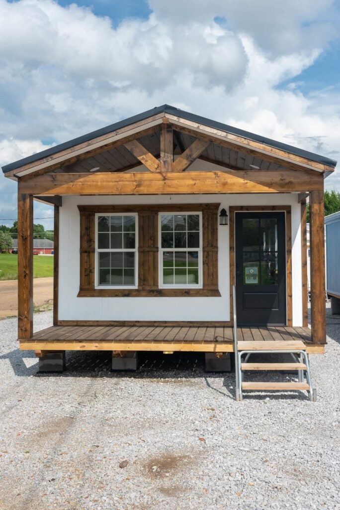 Small cabin with wooden accents and front porch steps, set on gravel. A black door and large windows add charm under a partly cloudy sky.