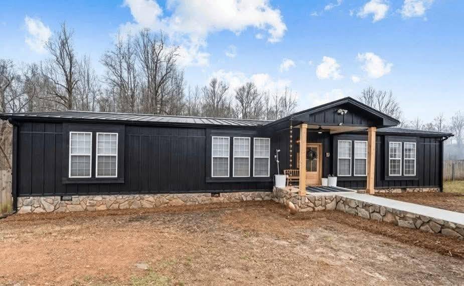 A single-story black house with stone accents, front porch, and wooden pillars. It has white framed windows, a clear blue sky, and leafless trees. Cozy and inviting atmosphere.