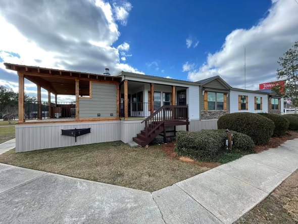 A modern single-story gray building with a wooden porch, stairs, and surrounding hedges. It is set against a clear blue sky with clouds, creating a welcoming tone.