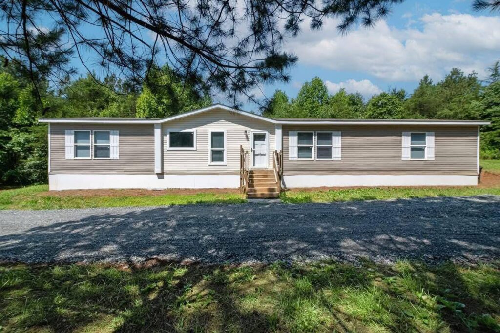 A light brown manufactured home with white shutters sits on a gravel driveway, surrounded by lush green trees under a bright blue sky.