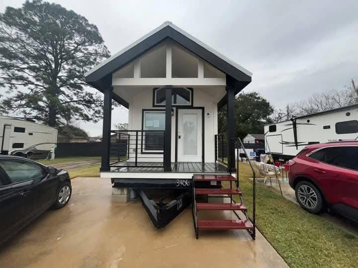 Compact tiny house with a modern design, featuring a black-and-white facade, front porch, and red steps, surrounded by parked cars and RVs.