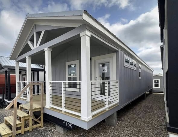 A modern tiny house with a blue-gray exterior and white trim features a small porch with white railings. The sky is partly cloudy, creating a serene ambiance.