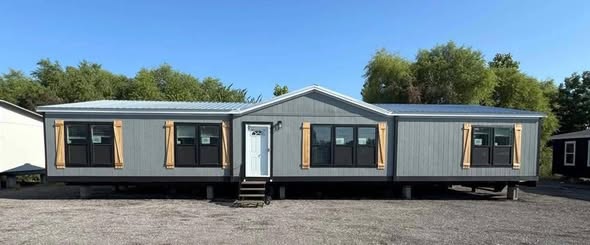 A wide gray mobile home with a white front door and multiple windows with wooden shutters stands on gravel. Trees and a clear blue sky are in the background.