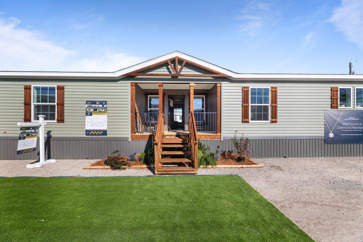 A modern manufactured home with a green lawn and wooden steps leading to a porch. The structure is light-colored with brown trim, conveying a welcoming and fresh atmosphere.