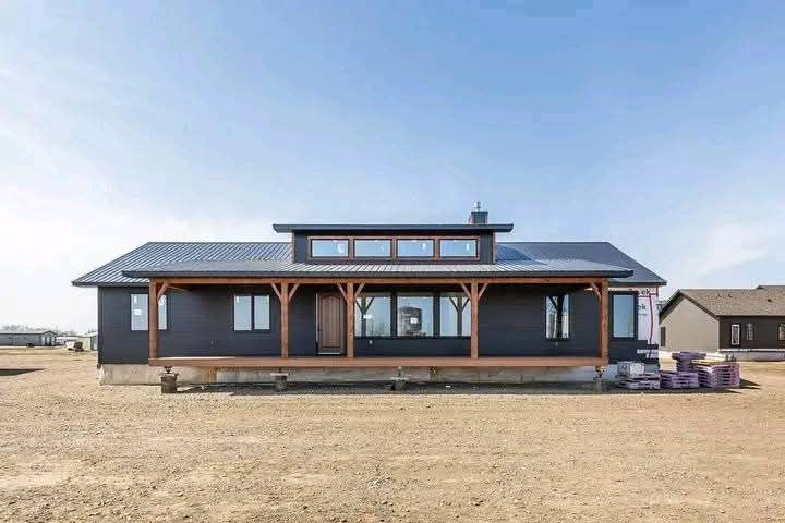 Single-story modern house with dark siding and large windows under a bright blue sky. The setting is a barren dirt area, conveying a sparse and tranquil atmosphere.