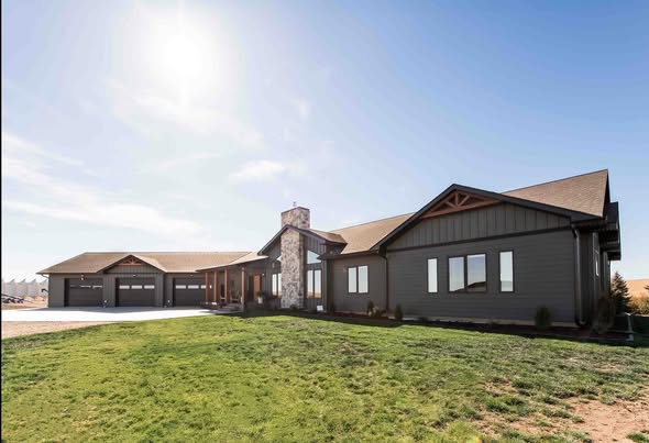 Modern single-story home under blue sky with stone chimney, large windows, and attached three-car garage. Sunlight creates a bright, welcoming feel.