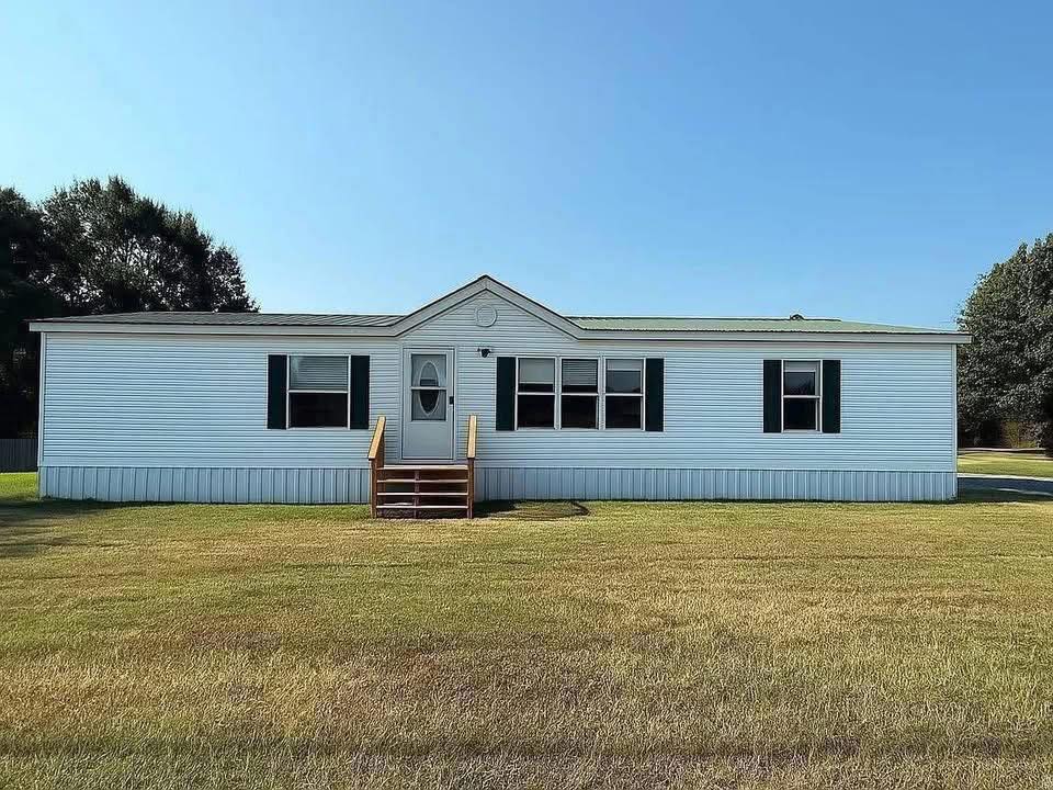 A white mobile home with green shutters sits on a grassy lawn under a clear blue sky. Wooden steps lead to the front door. Trees are in the background.
