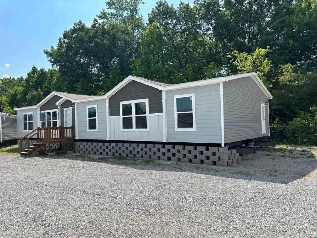 A modern gray mobile home on a gravel lot, elevated on concrete blocks, with white-trimmed windows and a small wooden porch. Surrounded by tall green trees under a clear blue sky.