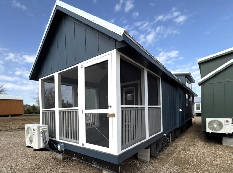 A charming tiny house with blue siding and a white-trimmed screened porch sits under a blue sky. An air conditioner unit is visible outside.