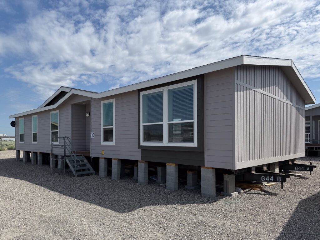 A gray modular home elevated on concrete blocks under a partly cloudy sky. The home features multiple windows and a small entry staircase, conveying a sense of simplicity and functionality.