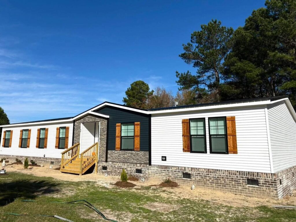 A spacious white and black modular home with wooden shutters stands under a clear blue sky, surrounded by trees and a simple yard with small plants.