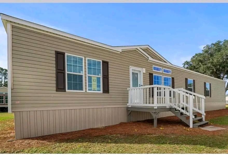 Single-story beige mobile home with white trim, a small front porch with stairs, and brown shutters. The sky is clear and a large tree is visible.
