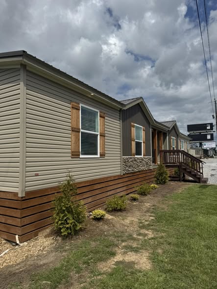 A modern manufactured home with beige siding and wooden shutters is shown on a cloudy day. Small shrubs are planted along the front, next to a neat lawn.