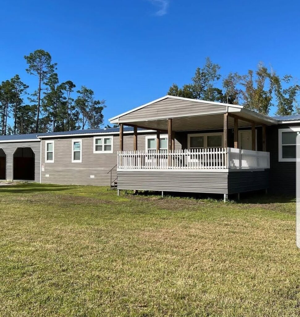 Single-story gray mobile home with a white-railed porch set on a grassy yard. Tall trees in the background under a clear blue sky. Tranquil setting.